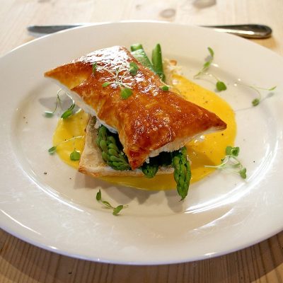 A golden puff pastry sits atop fresh asparagus spears and sauce on a white plate, garnished with microgreens, on a light wooden table. A fork and knife are in the background.