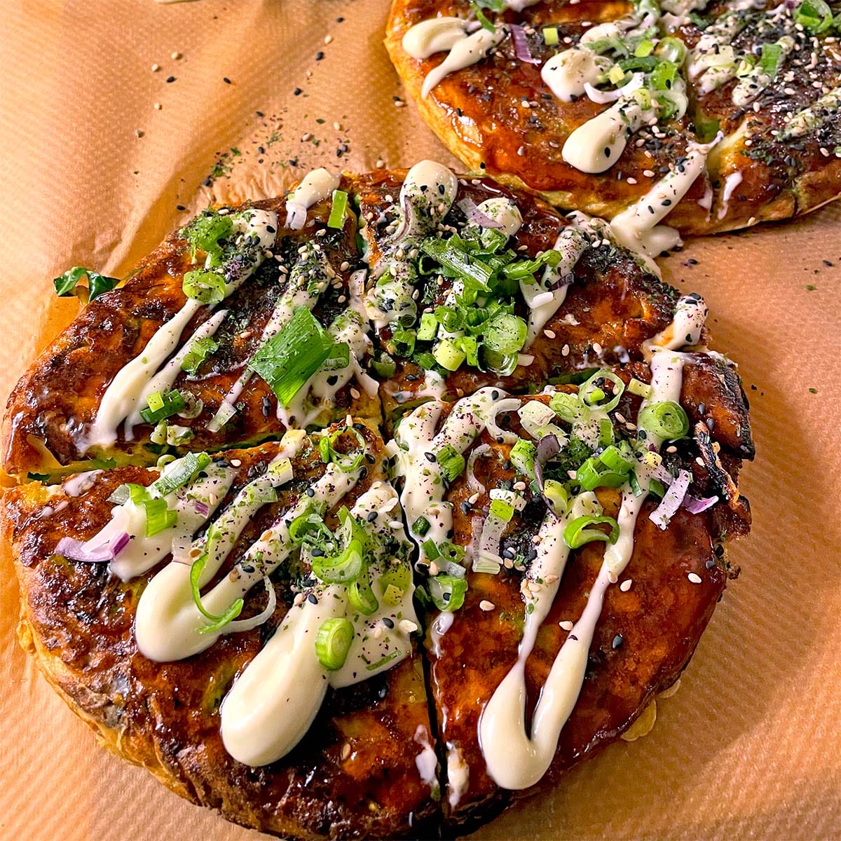 Two savoury Japanese pancakes (okonomiyaki) topped with mayonnaise, chopped spring onions, red onions, sesame seeds, and herbs, sitting on brown baking parchment.