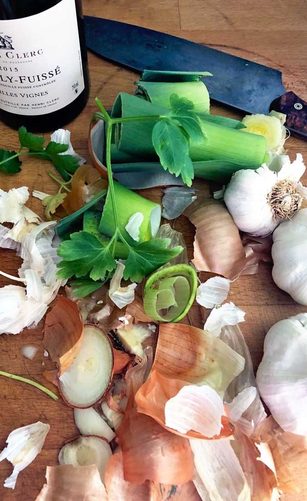 A chopping board with chopped leeks, parsley, garlic bulbs, onion skins, a knife, and a bottle of white wine. The scene suggests meal preparation with fresh vegetables and wine.